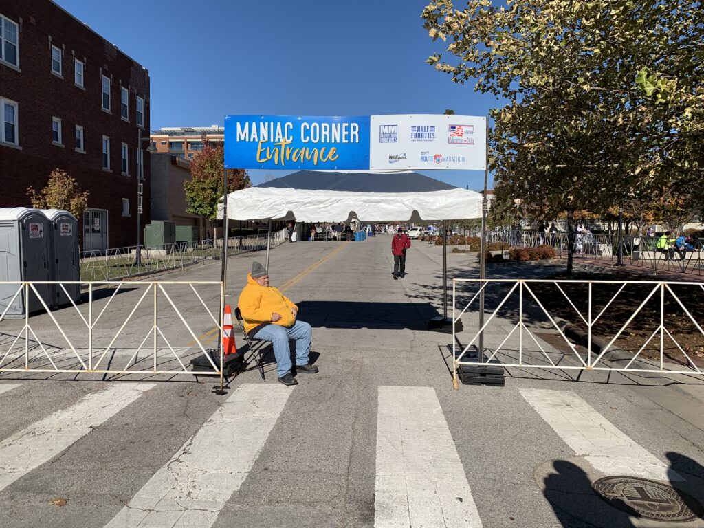 Man sitting beside Super H-Pole with Maniac Corner Entrance signage.