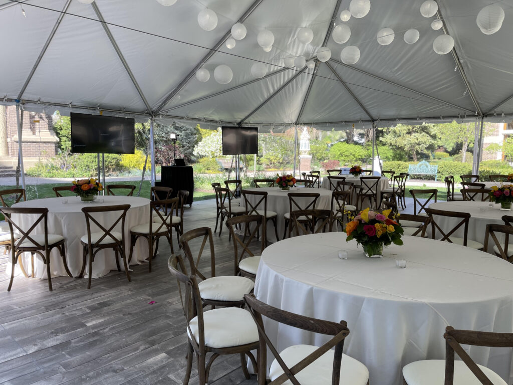 Tables and chairs with white linens set up under a large white outdoor tent with wood floors.