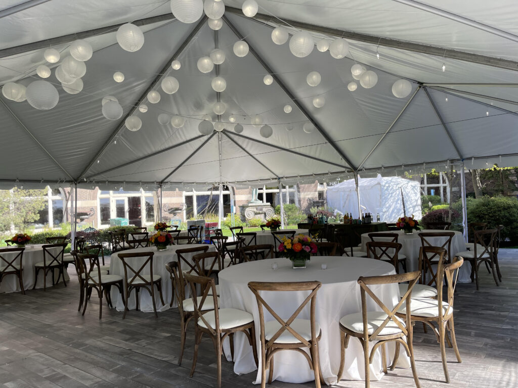 Tables and chairs with white linens set up under a large white outdoor tent with wood floors.