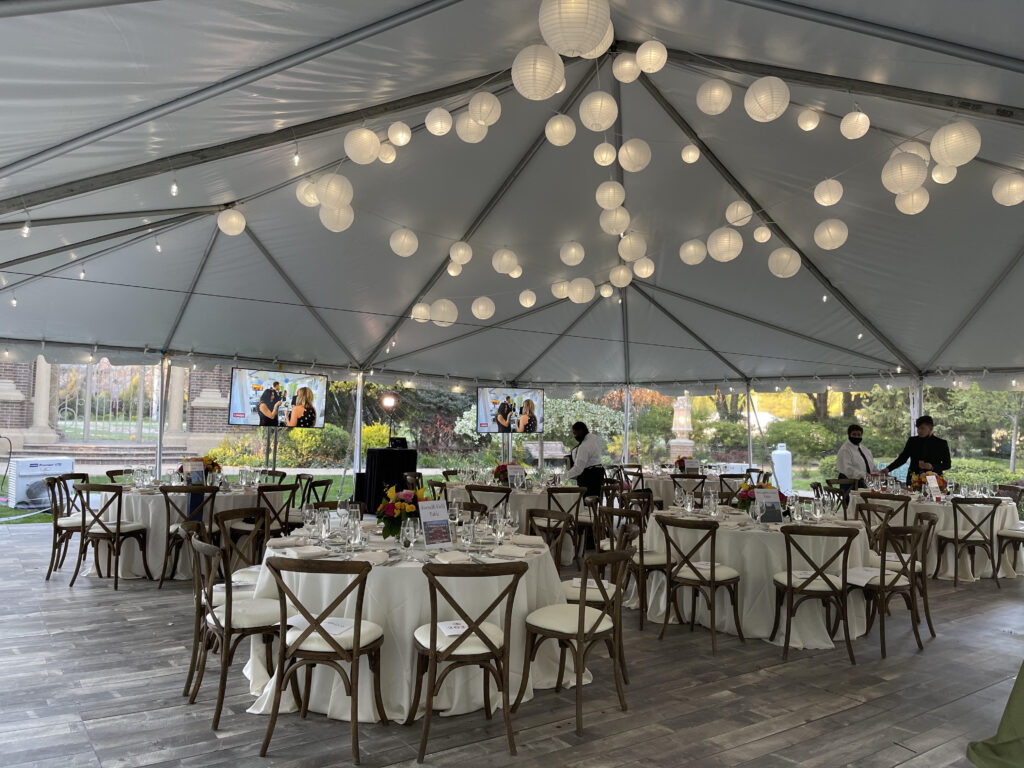 Tables and chairs with white linens set up under a large white outdoor tent with wood floors.